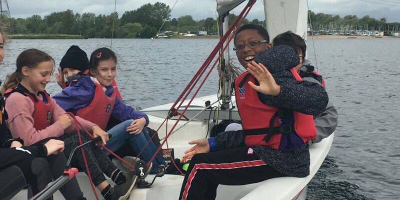 Young participants are in a sailboat on a calm day, whilst one of the sailors waves at the camera.